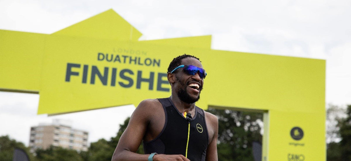 Athlète souriant en haut noir sans manches et lunettes de soleil bleues franchissant une arche d'arrivée jaune vif marquée DUATHLON FINISHES