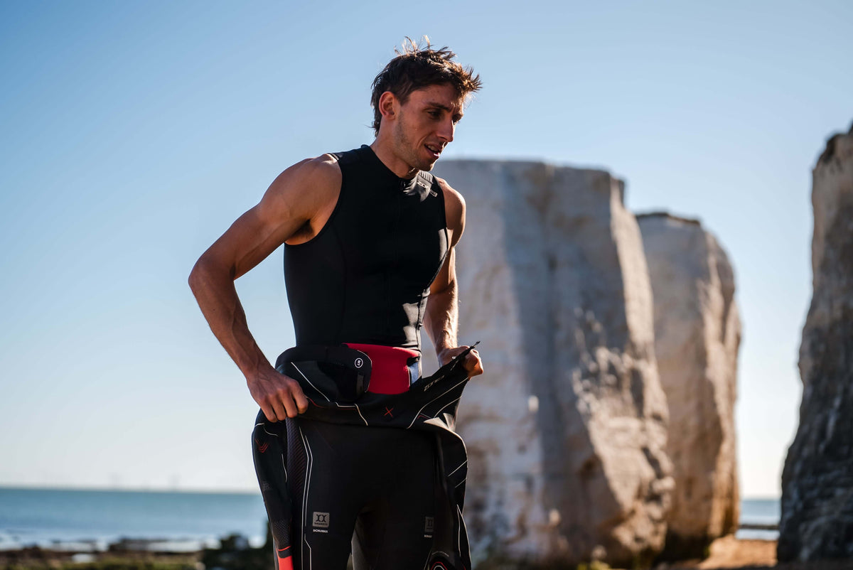 Athlète en combinaison néoprène noire et bonnet blanc debout près d'un rivage rocheux, un sac de transition étanche rouge à la taille.