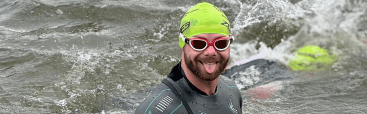 Triathlète avec bonnet de bain vert fluo et lunettes de natation rouges, souriant en nageant dans une eau agitée.