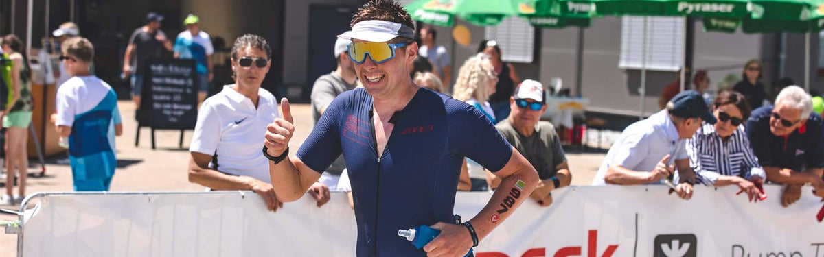 Triathlète en tenue bleu foncé avec visière blanche et lunettes de soleil miroir, souriant et levant le pouce devant des spectateurs derrière les barrières