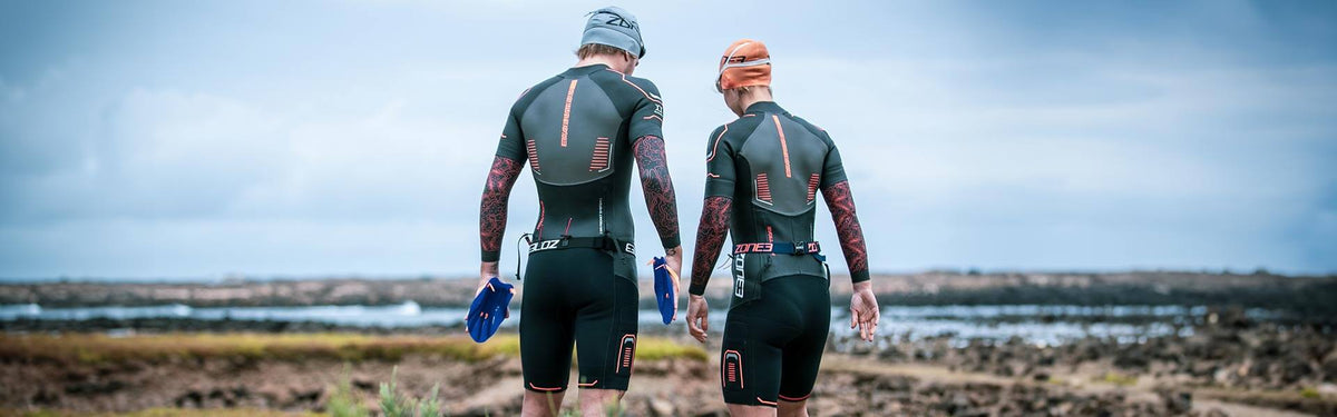 Deux athlètes en combinaisons néoprène Zone3 debout sur une côte rocheuse, des palmes de swimrun bleues à la main.