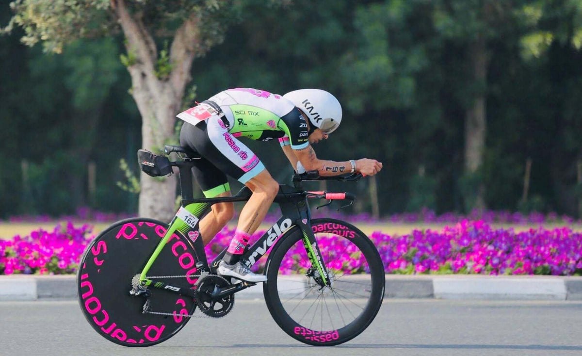 Triathlète en tenue vert fluo et rose sur un vélo de contre-la-montre, sur une route avec un parterre de fleurs en arrière-plan