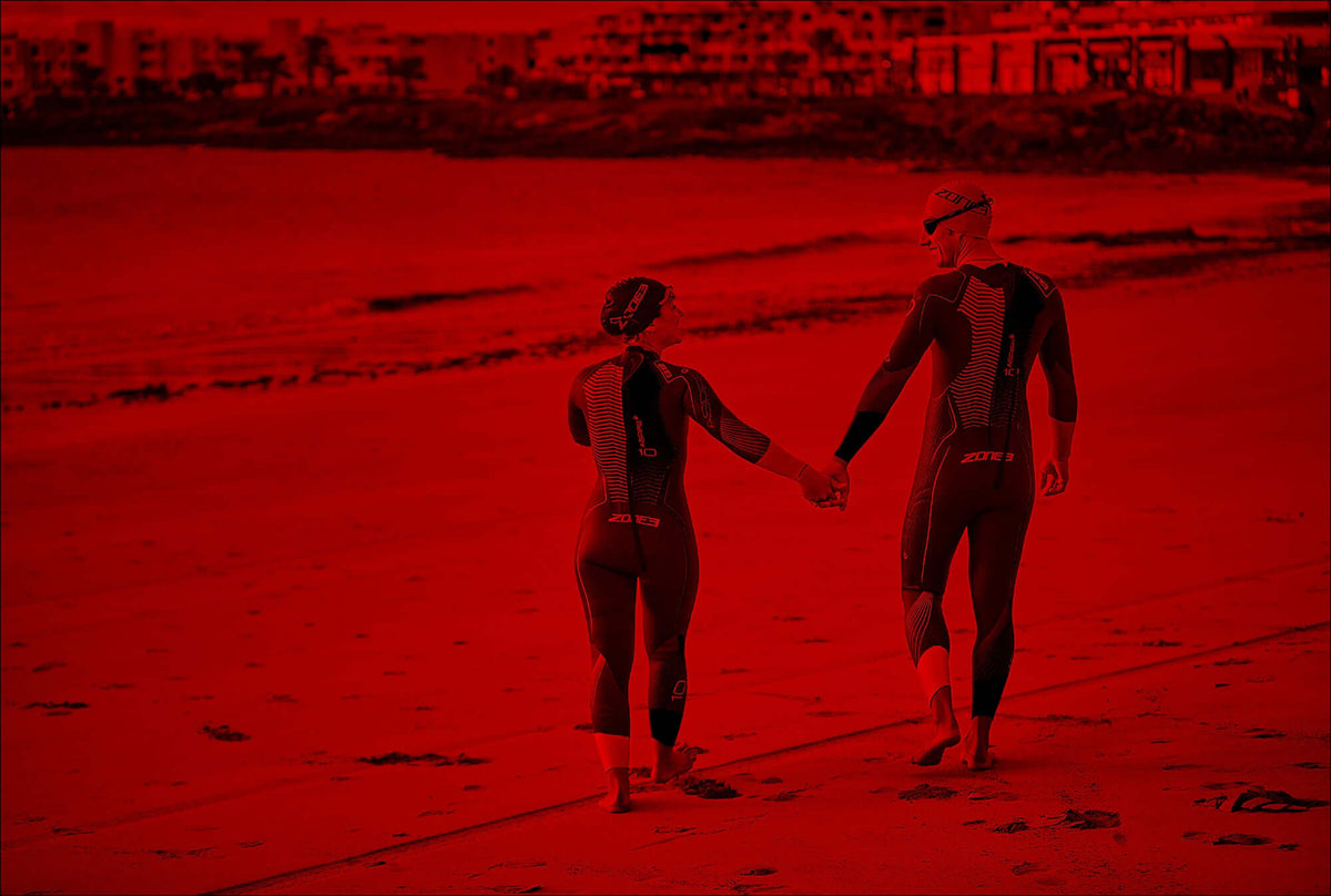 Deux athlètes en trifonctions assorties marchant main dans la main le long d'une plage au bord de l'eau