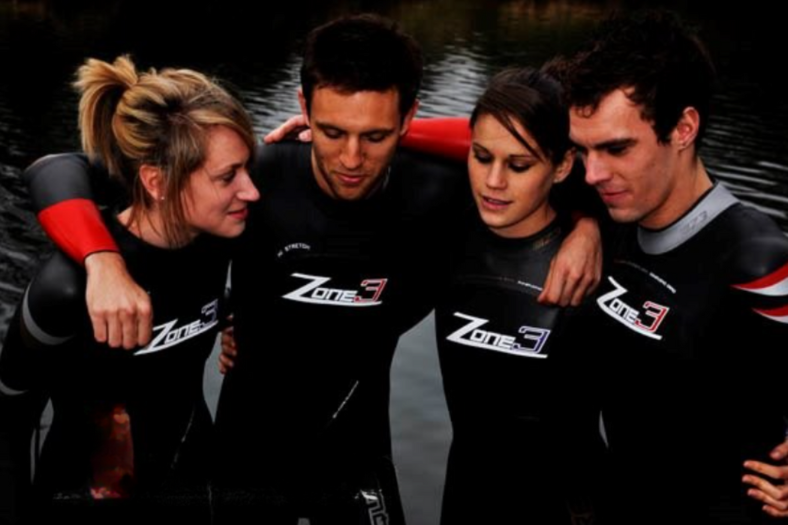 Photo de 4 personnes debout devant un plan d'eau, toutes vêtues de premières versions des combinaisons néoprène ZONE3. Deuxième en partant de la gauche : le fondateur James Lock.