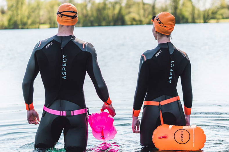 Deux personnes regardant un plan d'eau entouré d'arbres. Elles portent des combinaisons néoprène ZONE3 Aspect, des bonnets de bain orange et des lunettes de natation orange, ainsi que des bouées de natation ZONE3 attachées à la taille.