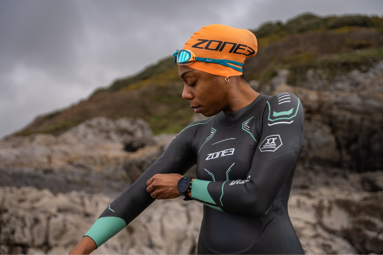 Une femme portant une combinaison néoprène ZONE3 Vision devant un décor rocheux. Elle porte un bonnet de bain ZONE3 orange et des lunettes de natation bleu canard, et ajuste le bras droit de sa combinaison néoprène.