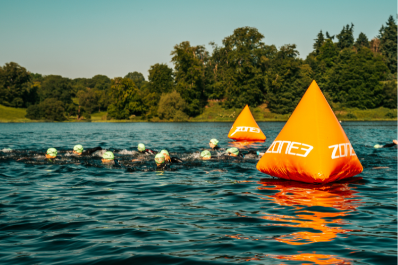 Un plan d'eau libre entouré d'arbres et de verdure. Au milieu de l'eau, des nageurs et deux bouées orange en forme de pyramide avec le logo ZONE3.