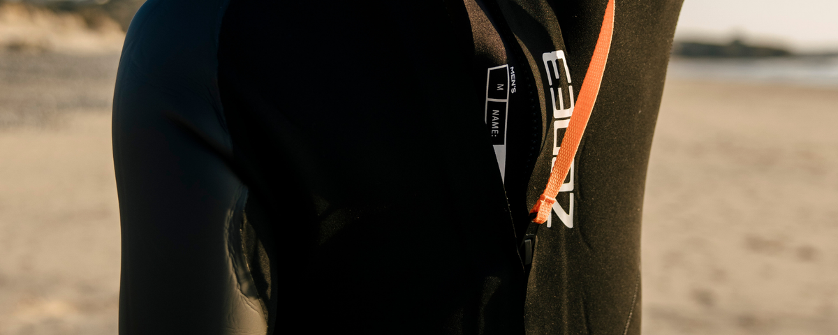 Close-up of a person wearing a black wetsuit with orange accents on a beach.