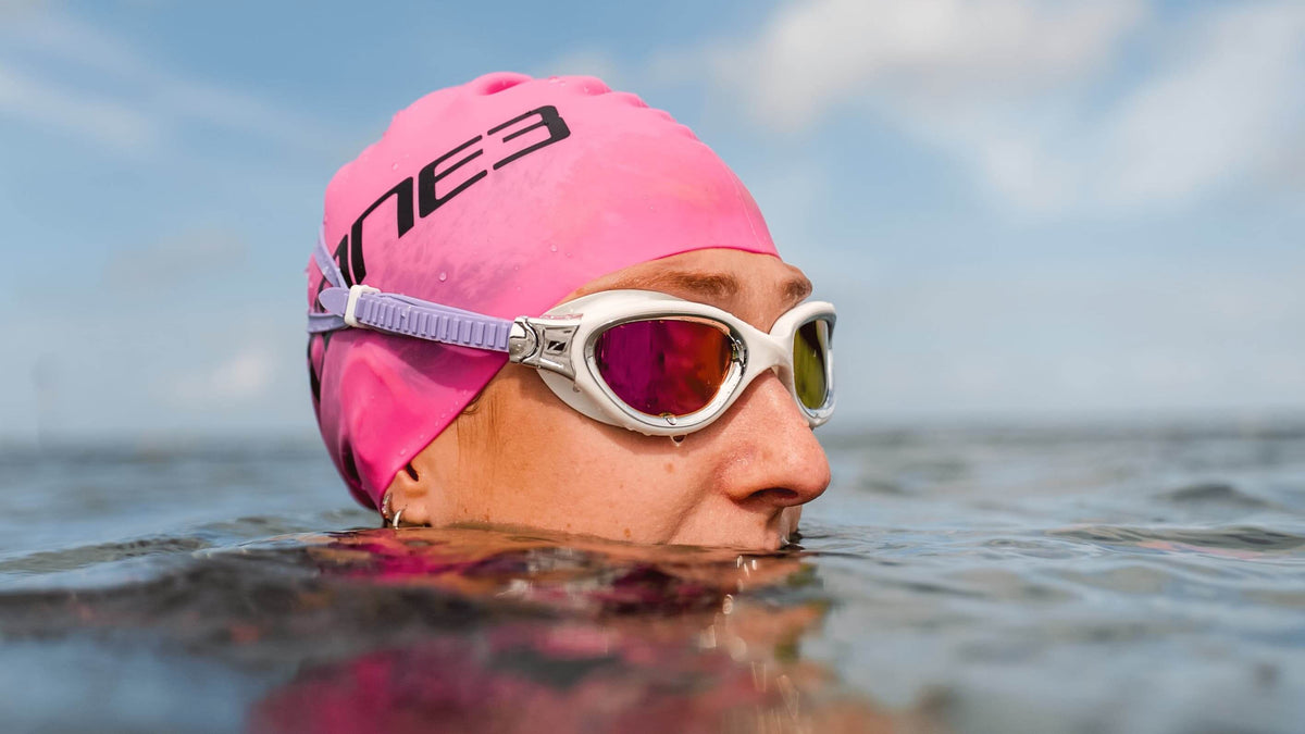Athlete swimming in open water wearing Zone3 pink swim cap and Zone3 white/lilac swim goggles