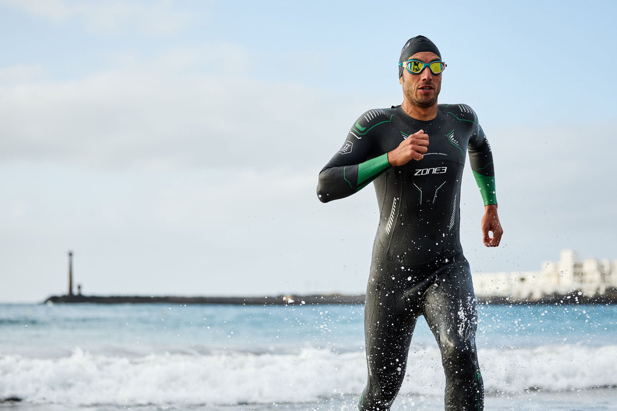 Person in a wetsuit and swim cap standing on a beach with ocean waves and sky in the background